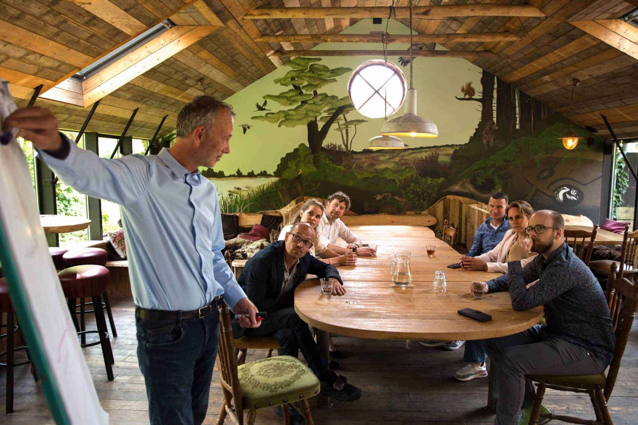 Een groep mensen vergadert in een zaal met natuurlijke element. Man geeft presentatie op een flip-over